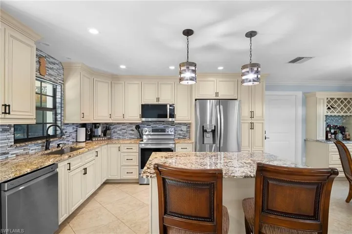 Kitchen with cream cabinets, stainless steel appliances, backsplash, hanging light fixtures, and light stone countertops