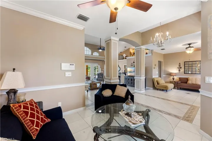 Living room featuring light tile patterned flooring, ceiling fan with notable chandelier, crown molding, and decorative columns