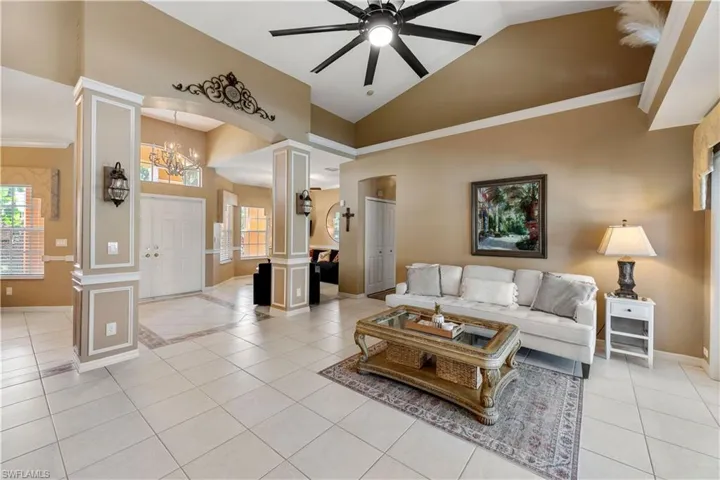 Tiled living room with decorative columns, ceiling fan with notable chandelier, crown molding, and high vaulted ceiling