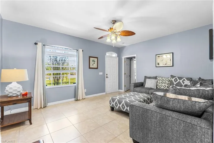 Living area featuring neutral tile flooring, a window with horizontal blinds and drapes, and a ceiling fan with light fixture