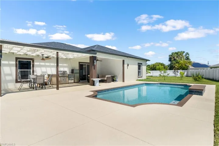 Outdoor living space featuring a swimming pool with integrated seating, extensive concrete patio, and a covered lanai with an outdoor kitchen setup