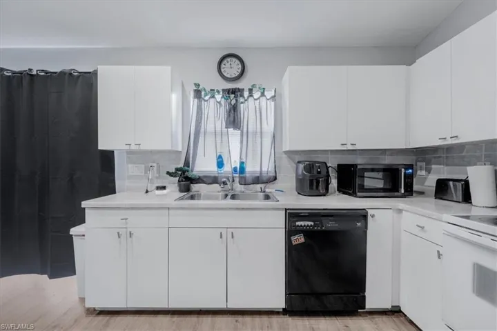 Kitchen featuring white cabinetry, light-toned countertops, a stainless steel double sink, a dark tile backsplash, and wood-finish flooring