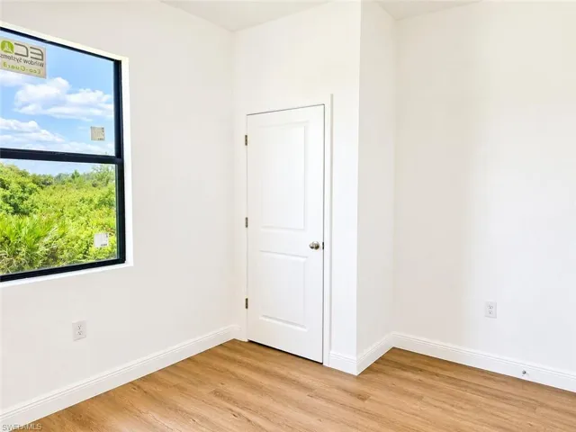 Bright interior space featuring a large window with black framing, wood-finish flooring, white baseboards, and a white two-panel door with a brushed nickel knob