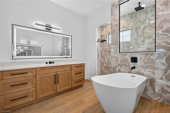 Bathroom featuring a freestanding soaking tub, large vanity with light wood-finish cabinetry, stone-look tile accents, and modern black fixtures