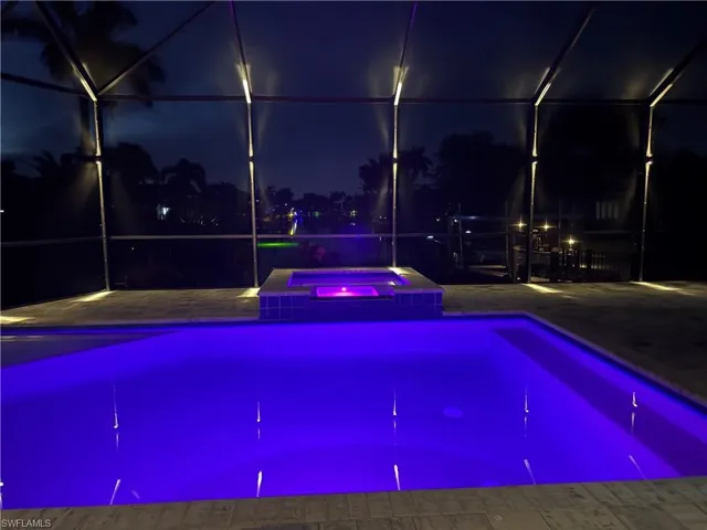 Screened pool enclosure featuring a pool with vibrant blue underwater lighting, an integrated hot tub with purple lighting, and patterned paver decking