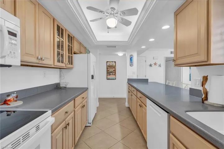 Kitchen with a raised ceiling, crown molding, white appliances, light brown cabinetry, and light tile patterned floors