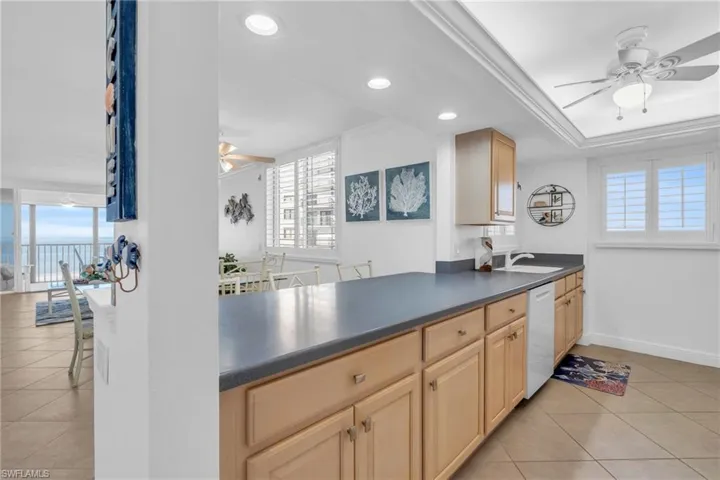 Kitchen with light brown cabinetry, plenty of natural light, white dishwasher, and light tile patterned floors, virtually enhanced image