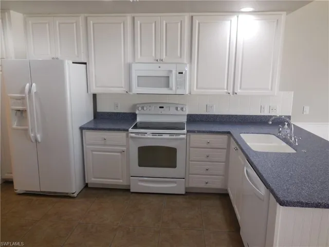 Kitchen with tasteful backsplash, white cabinets, dark tile patterned flooring, white appliances, and sink