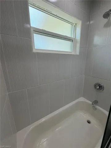 White bathtub with chrome fixtures, surrounded by light gray vertical tile with a subtle textured finish, and a frosted window