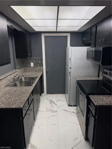 Galley kitchen with dark cabinetry, granite-finish countertops, stainless steel double basin sink, and white marbling-effect tile flooring