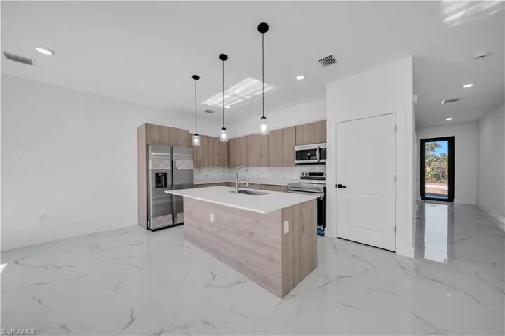 Kitchen featuring modern cabinets, stainless steel appliances, an island with sink, decorative light fixtures, and light stone countertops