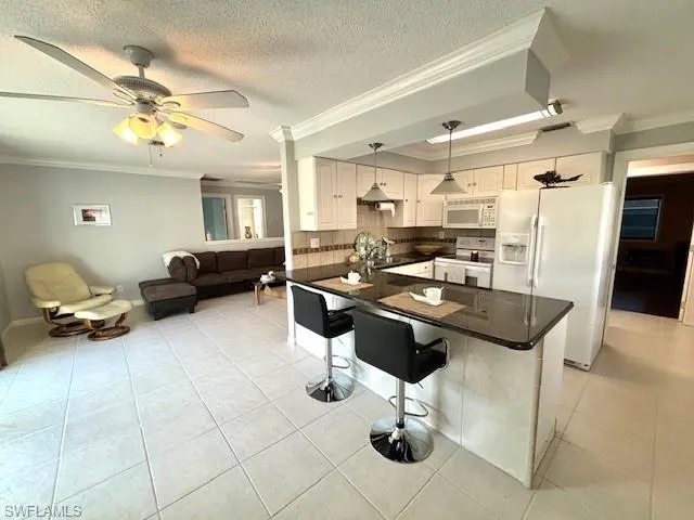 Kitchen with hanging light fixtures, a kitchen bar, white cabinetry, ornamental molding, and white appliances