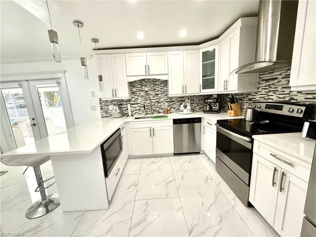 Kitchen featuring stainless steel appliances, pendant lighting, white cabinets, light marble finish flooring, and glass fronted cabinets