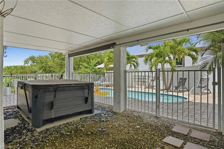Covered patio featuring a hot tub on a pebble base, opening to a fenced pool area with palm trees and a paved deck - Virtually Edited Image