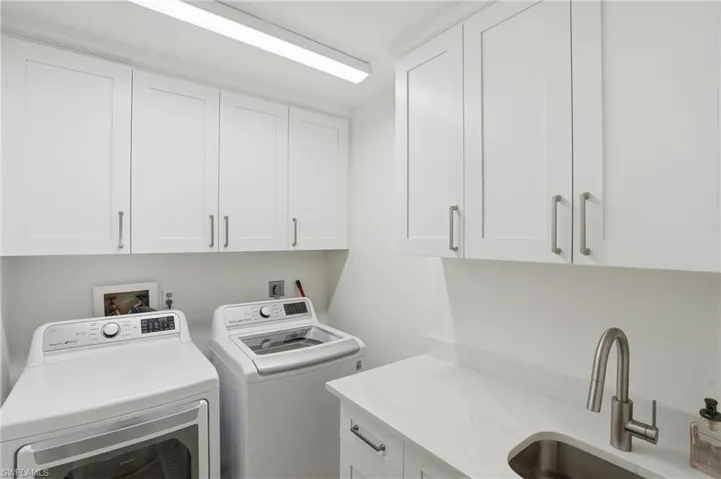 Laundry area featuring white shaker-style cabinetry, a white countertop with an integrated sink, a brushed nickel faucet, and overhead linear lighting - Virtually Edited Image