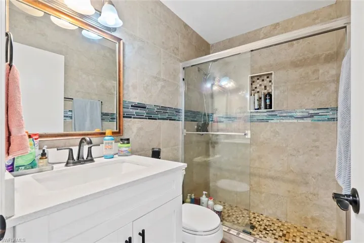 White vanity with a quartz countertop and integrated sink, framed mirror with overhead lighting, tiled shower with a glass enclosure, decorative tile accent band, and a mosaic tile shower floor