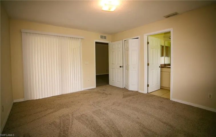 This room features a light-colored carpet, vertical blinds on a large window, a ceiling light fixture, and a neutral wall color