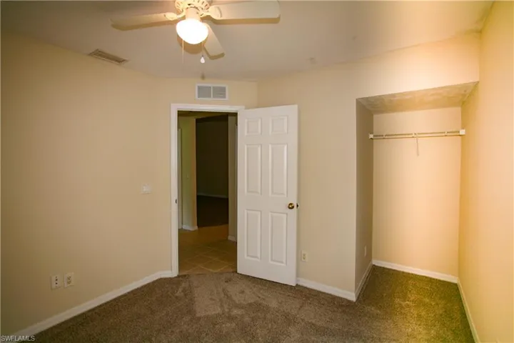 The room features neutral-toned walls, carpeted flooring, a ceiling fan with an integrated light fixture, and an open closet space with a clothing rod