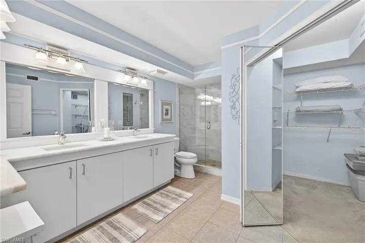 Primary Bathroom Dual vanity with white cabinetry and integrated sinks, a glass-enclosed shower with tile surround, a built-in toilet, and an expansive walk-in closet with wire shelving and a mirrored sliding door