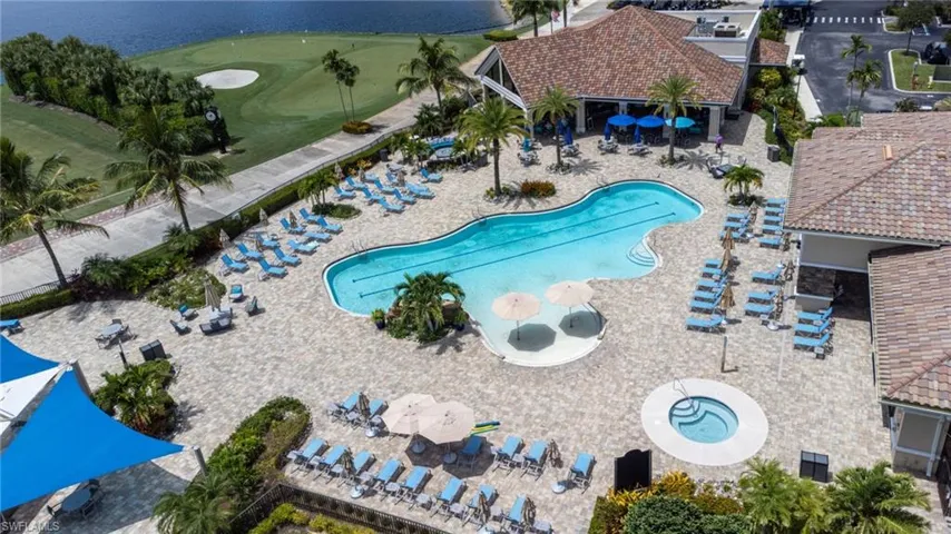 Resort-style pool with integrated spa, expansive paver deck, adjacent clubhouse with tiled roof, lush tropical landscaping, and a golf course with water features