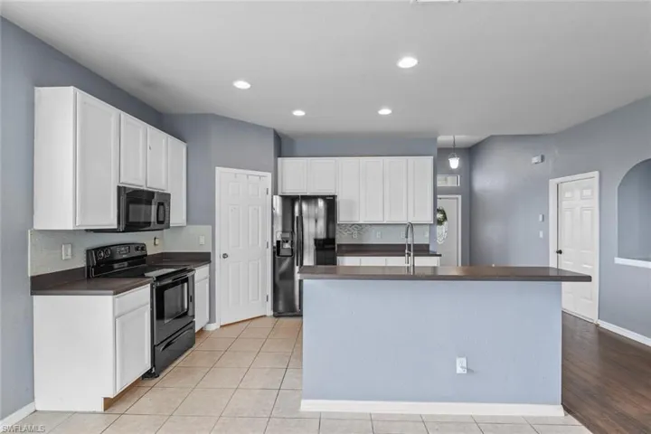 Kitchen with dark countertops, backsplash, black appliances, a kitchen island with sink, and white cabinets