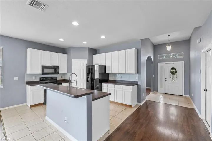 Kitchen with arched walkways, black appliances, a kitchen island with sink, white cabinets, and light tile patterned floors