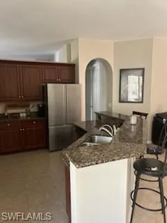 Kitchen featuring a kitchen bar, freestanding refrigerator, dark stone counters, arched walkways, and a center island with sink