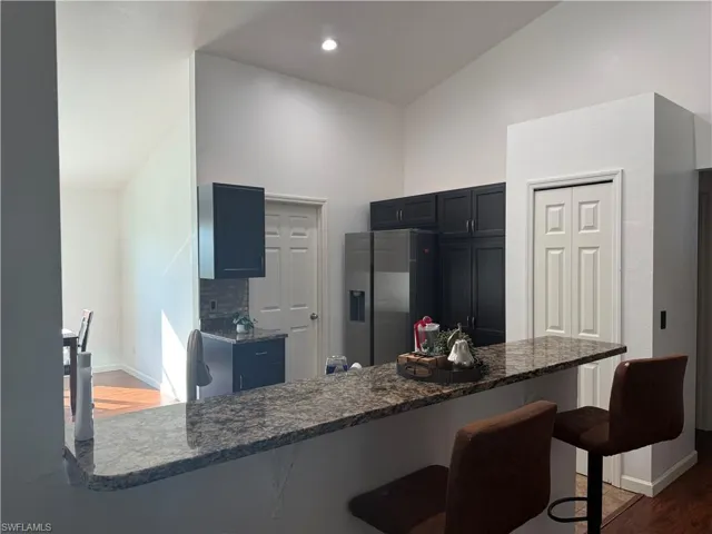 Kitchen view from living room featuring vaulted ceiling, dark wood-type flooring, a kitchen bar, stainless steel fridge with ice dispenser, and dark stone counters
