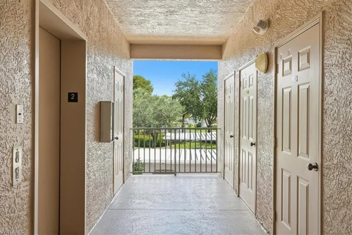 Outdoor corridor with textured walls, multiple panel doors, an elevator, and a balcony with metal railings
