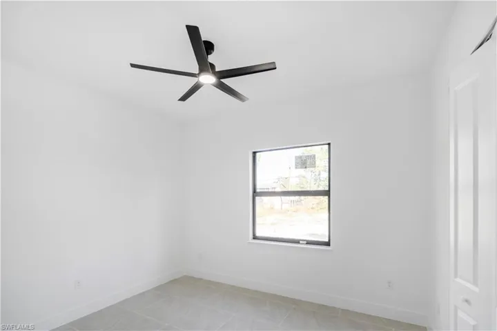 Bright interior room featuring a modern black ceiling fan with integrated lighting, a single pane window with dark trim, light-toned tile flooring, white walls, and white baseboards