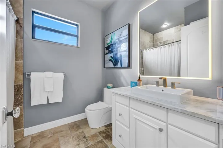 Bathroom featuring a white vanity cabinet with a rectangular vessel sink, stone-surface countertop, LED-backlit mirror, and neutral-toned tiled flooring