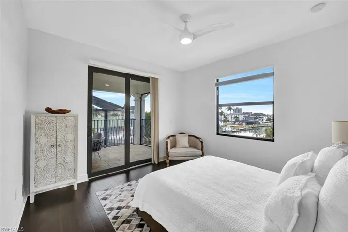 Dark wood-finish flooring, sliding glass door with access to a covered balcony, ceiling fan with integrated lighting, and expansive window with water backdrop