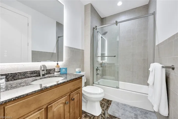Bathroom featuring a stone-topped vanity with an undermount sink, illuminated mirror, shower-tub combination with sliding glass doors, and tiled wall finishes