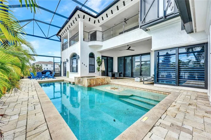 Screened-in pool area featuring a raised spillover spa, stone-paver decking, a two-story balcony with metal railings, and sliding glass wall panels