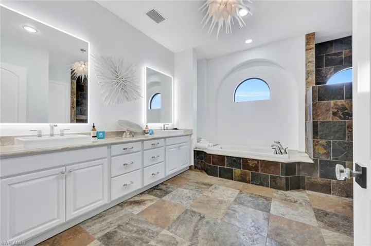 Dual-vanity bathroom featuring a stone-tile floor and surround, drop-in soaking tub within an arched alcove, backlit mirrors, and extensive white cabinetry
