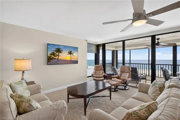 Carpeted living area with a ceiling fan, wall-mounted television, and floor-to-ceiling windows accessing a balcony overlooking the ocean