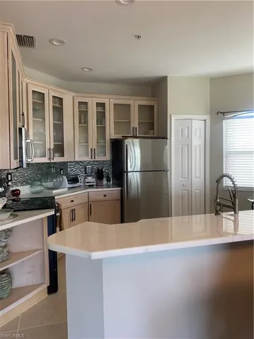 Kitchen with light brown cabinetry, appliances with stainless steel finishes, decorative backsplash, glass insert cabinets, and light stone counters