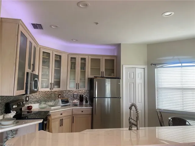 Kitchen featuring stainless steel appliances, light brown cabinets, light stone counters, glass insert cabinets, and tasteful backsplash
