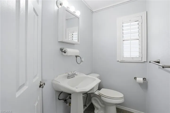 Pedestal sink with chrome faucet, wall-mounted medicine cabinet with integrated lighting, window with white plantation shutters, and light blue wall paneling