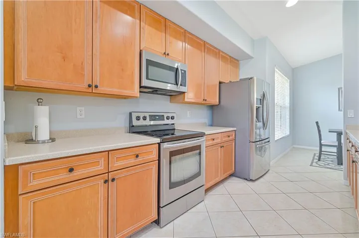 Kitchen with stainless steel appliances, light tile patterned flooring, lofted ceiling, light wood finish cabinets, and light stone counters