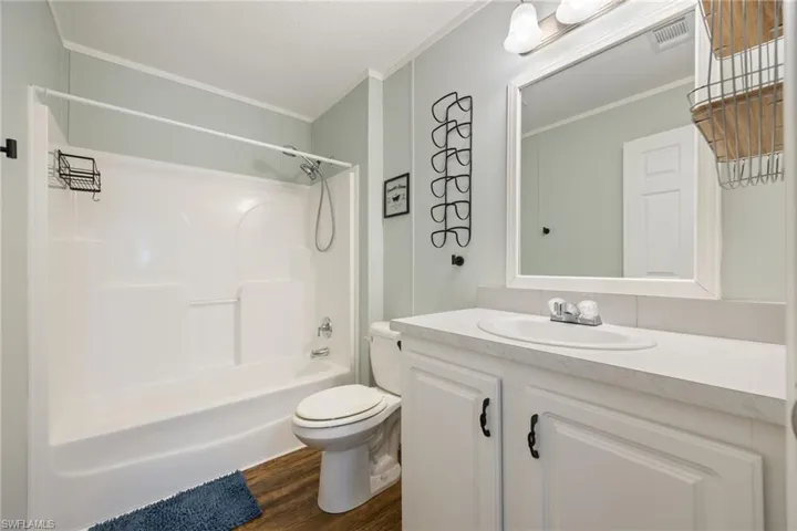 Guest bathroom featuring a white integrated tub and shower unit, a white vanity with an integrated sink and base cabinetry, wood-finish flooring, a wall-mounted mirror, and a two-light vanity fixture.