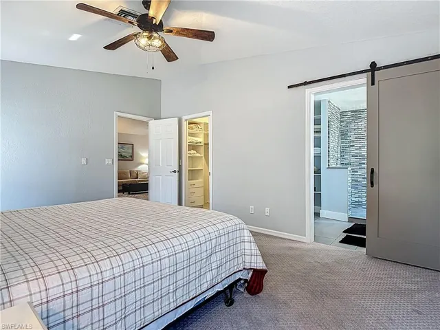 Bedroom featuring vaulted ceiling, a barn door, ensuite bath, light colored carpet, and a ceiling fan