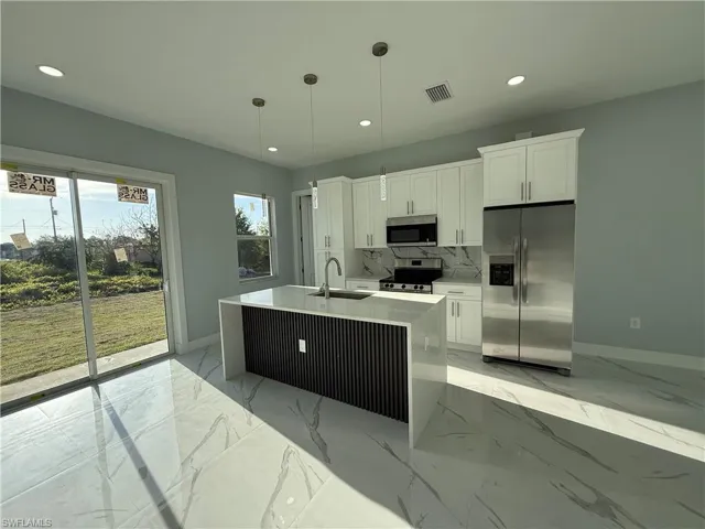 Kitchen with stainless steel appliances, light stone counters, a kitchen island with sink, white cabinets, and decorative light fixtures