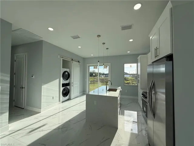 Kitchen featuring light stone counters, a barn door, an island with sink, stainless steel refrigerator with ice dispenser, and stacked washer and clothes dryer