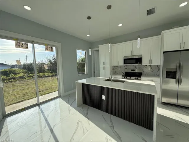 Kitchen with light stone counters, stainless steel appliances, an island with sink, white cabinets, and decorative light fixtures