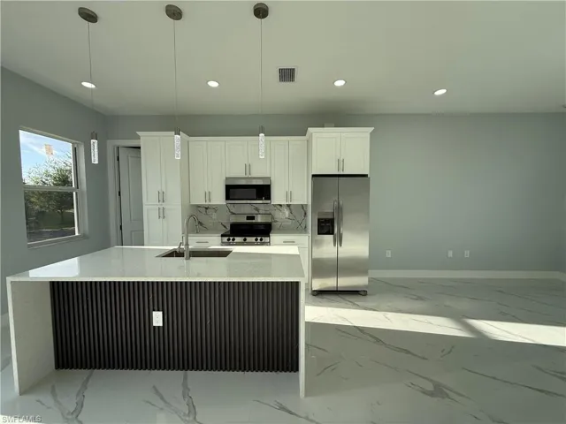Kitchen featuring light stone countertops, stainless steel appliances, white cabinetry, a center island with sink, and decorative light fixtures