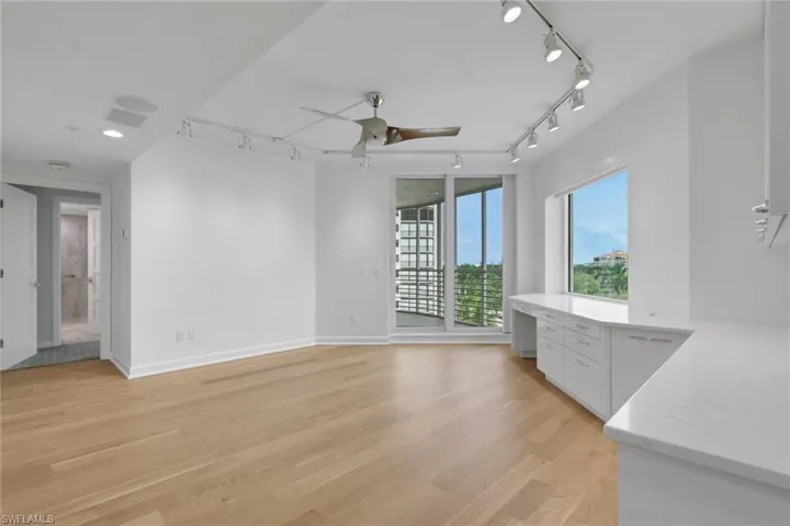 Bright, open living space featuring wide-plank wood-finish flooring, floor-to-ceiling windows with natural light, track lighting, ceiling fan, and a built-in white cabinetry with countertop workspace