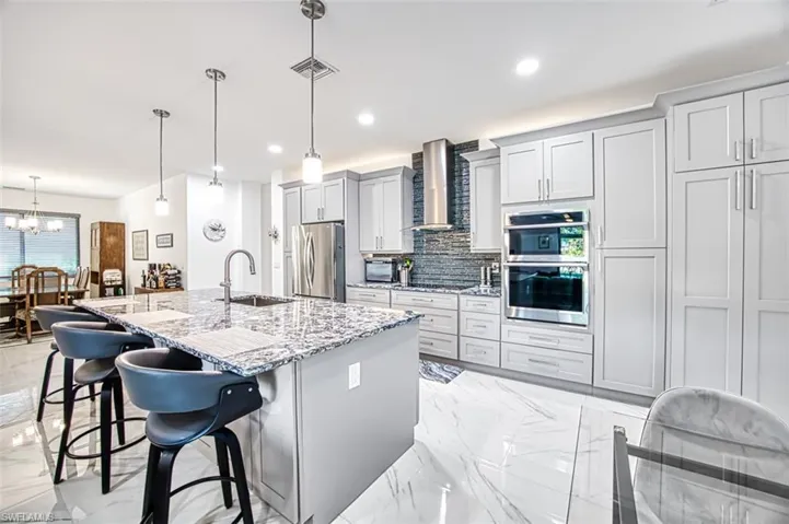 Kitchen with stainless steel appliances, light tile floors, hanging light fixtures, wall chimney exhaust hood, and a kitchen island with sink