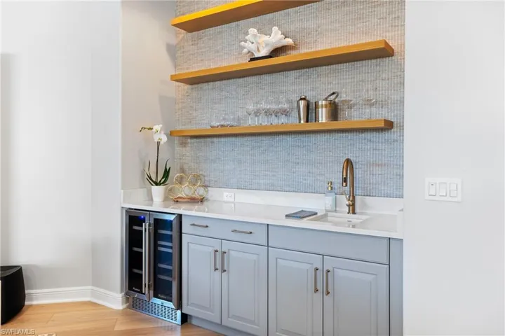 Bar featuring sink, light blue cabinetry, beverage cooler, decorative backsplash, gold hardware and floating shelves