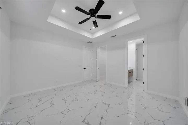 Unfurnished bedroom featuring recessed lighting, ceiling fan, ensuite bathroom, light marble finish floors, and a raised ceiling